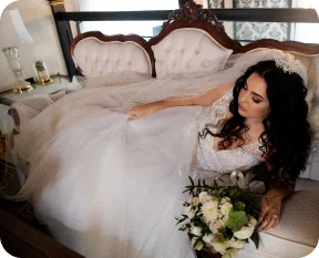 Bride in a white gown relaxing on a vintage sofa, holding a bouquet of white flowers.